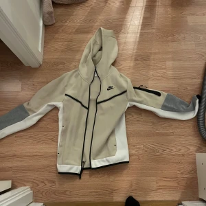 Beige och grå hoodie från Nike. (Nike tech)  - Snygg hoodie från Nike i beige och grått med svarta detaljer. Hoodie har en dragkedja framtill och en praktisk huva. Perfekt för en sportig look. Nike tech 