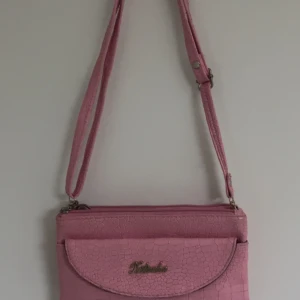 Rosa axelväska från Xilusha - Snygg rosa axelväska från Xilusha med krokodilpräglat mönster. Väskan har en justerbar axelrem och en praktisk dragkedja baktill. Perfekt för att ge en färgklick till din outfit!