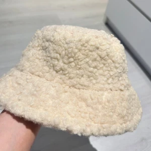 Beige teddyhatt - Mysig beige hatt i teddy-material, perfekt för kyliga dagar. Den har en mjuk och fluffig textur som ger en trendig look. Helt ny!