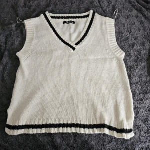 Vit stickad väst med svarta detaljer - Snygg vit stickad väst med svarta ränder vid v-ringningen och nederkanten. Perfekt för lager-på-lager och ger en stilren look.