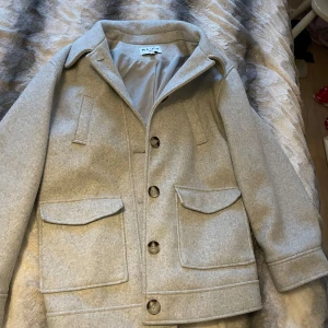 Grå kappa från NA-KD - Säljer en stilren beige kappa från NA-KD. Jackan har en klassisk design med knappar framtill och stora fickor. Perfekt för höst och vår, med en bekväm passform som ger en snygg look.