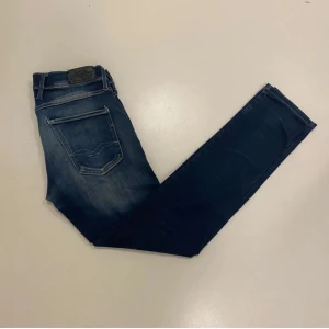 Replay anbass jeans med slitningar - Snygga blå jeans från Replay i modellen Anbass Hyperflex (slim fit) med slitna detaljer och en klassisk femficksdesign. De har en dragkedja och knapp framtill samt Replay-logga på bakfickan. Perfekta för en avslappnad stil.