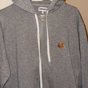 Grå hoodie från Kenzo - Säljer en stilren grå hoodie från Kenzo med dragkedja och en broderad tigerlogga på bröstet. Tröjan har en justerbar huva med vita snören och är perfekt för en avslappnad look. Pris kan diskuteras💯💯