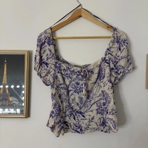 Blommig kortärmad blus - Charmig blommig blus i beige och blått med korta ärmar och knytning framtill. Perfekt för en somrig look med sitt luftiga och lätta material.