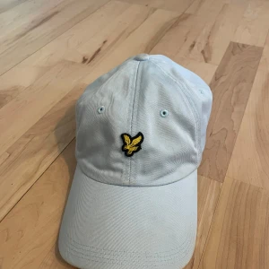 Ljusblå keps från Lyle & Scott - Snygg ljusblå keps från Lyle & Scott med en broderad gul logotyp framtill. Kepsen har en justerbar rem baktill för perfekt passform. Skriv privat om ni har frågor eller funderingar!