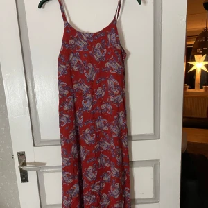 Röd mönstrad klänning med smala axelband - Snygg röd klänning med paisleymönster i blått och vitt. Den har smala axelband och en avslappnad passform, perfekt för varma dagar. Golvlång, snörning i midjan. One sized