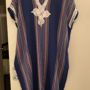 Blå randig klänning med dekorativt mönster - Säljer en unik blå klänning med färgglada ränder och ett dekorativt vitt mönster framtill. Klänningen har korta ärmar och en avslappnad passform. Perfekt för en bohemisk stil.