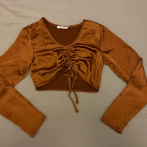 Brun croppad topp  - Snygg brun croppad topp i glansigt material. Toppen har långa ärmar och en v-ringad design med knytning framtill för en justerbar passform.