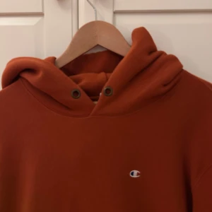 Hoodie från Champion - Säljer en snygg orange hoodie från Champion med klassisk logga på bröstet och ärmen. Tröjan har en stor ficka framtill och justerbar huva med snören. Perfekt för en avslappnad stil.