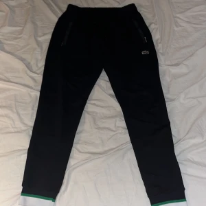 Svarta mjukisbyxor från Lacoste - Snygga svarta mjukisbyxor från Lacoste med elastisk midja och dragkedjefickor. Byxorna har gröna och vita detaljer vid bensluten och den klassiska krokodilloggan på sidan. Perfekta för en avslappnad stil.