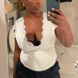 Vit spetsblus - Säljer en vacker vit spetsblus med djup v-ringning och fina spetsdetaljer. Blusen har en omlott-design och är ärmlös, vilket ger en elegant look. Perfekt för en somrig dag eller en speciell kväll.