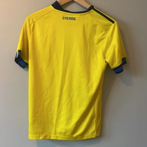 Gul Sverige fotbollströja från Adidas - Säljer en gul Sverige fotbollströja från Adidas med blå detaljer och korta ärmar. Tröjan har Sveriges emblem på bröstet och är tillverkad i AEROREADY-material för optimal komfort. Perfekt för fotbollsfans!