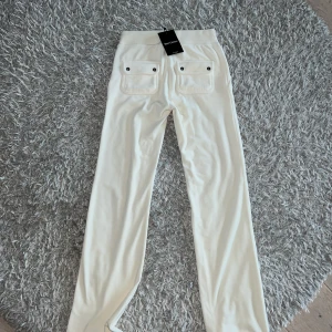 Vita mjukisbyxor från Juicy Couture - Snygga vita mjukisbyxor från Juicy Couture med broderad logga på fickan. Byxorna har en bekväm passform med dragsko i midjan och två bakfickor med knappar. Perfekta för en avslappnad stil.