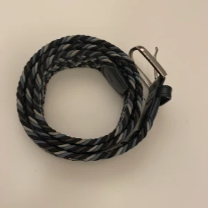 Flätat bälte från John Henric - Snyggt flätat bälte från John Henric i olika nyanser av svart, grått och blått. Bältet har en klassisk metallspänne och är perfekt för att ge en stilren touch till din outfit.