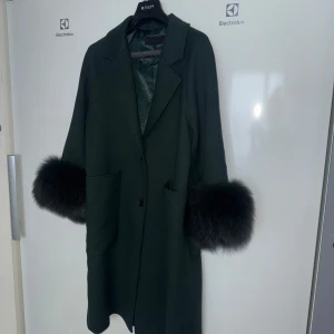 Grön kappa med pälsdetaljer - Elegant grön kappa med långa ärmar och stiliga pälsdetaljer vid ärmsluten. Perfekt för att hålla sig varm och snygg under kyliga dagar. Kappan har en klassisk design med knappar framtill och praktiska fickor.