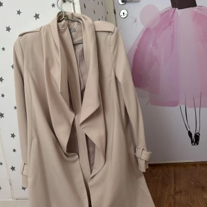 Beige trenchcoat från H&M - Snygg beige trenchcoat från H&M med knytband i midjan och stilrena detaljer. Jackan har långa ärmar med justerbara remmar och en klassisk krage. Perfekt för en chic look.