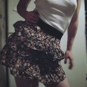 Blommig volangkjol - Charmig kortkjol med blommigt mönster i rosa och grönt på en svart bakgrund. Kjolen har flera lager av volanger som ger en lekfull look. Perfekt för att addera en romantisk touch till din outfit.