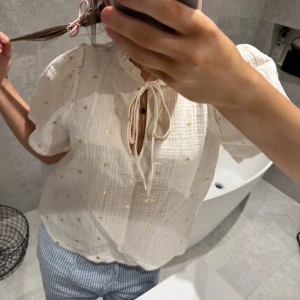 Vit blus från & Other Stories - Säljer en söt cremevit blus från & Other Stories med små guldhjärtan och knytning framtill. Blusen har korta puffärmar och en luftig passform. Perfekt för en somrig look! 🌼