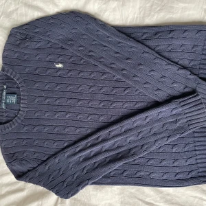 Mörkblå kabelstickad tröja från Ralph Lauren - Säljer en snygg mörkblå stickad tröja från Ralph Lauren med kabelstickat mönster. Den är använd men väl omhändertagen. Det finns tecken på slitning och färgen är aningen ljusare runt kragen på tröjan och på ett ställe på armen (se sista bilden). Tröjan är lite mörkare än hur den ser ut i bilderna💗nypris 1990kr