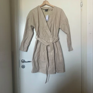 Beige trenchcoat med bälte - Snygg beige klänning från Gina Tricot med knytbälte i midjan. Klänningen har en stilren design med långa ärmar och är perfekt för vår och höst. Skinn imitation. Den är tillverkad i ett syntetmaterial som ger en modern look. Storlek XS men passar en XS/S. Helt ny med tags. 