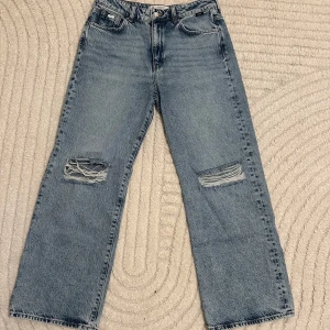 Blå jeans från Mavi - Säljer ett par blå jeans från Mavi med slitna detaljer på knäna. De har en klassisk femficksdesign och knappgylf. Perfekta för en avslappnad stil.