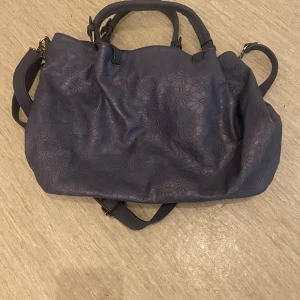 Blå axelväska med blommigt foder - Snygg blå axelväska med blommigt mönster på insidan. Väskan har både handtag och en justerbar axelrem för flexibilitet. Utsidan är dekorerad med ett subtilt blommönster och har en praktisk dragkedja på baksidan. Perfekt för att bära med sig allt man behöver.