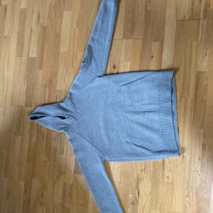 Grå hoodie från Stone Island - Säljer en stilren grå hoodie från Stone Island med deras ikoniska logga på ärmen. Tröjan har en praktisk magficka och är tillverkad i ett mjukt material som passar perfekt för kyligare dagar. Perfekt för dig som vill ha både stil och komfort.