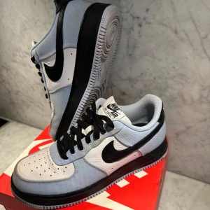 Nike Air Force i blått och vitt - Snygga Nike Air Force sneakers i en fräsch kombination av blått och vitt med svarta detaljer. Skorna har en klassisk design med snörning och en vit sula. Perfekta för en stilren look. Ny pris: runt 2000 kr