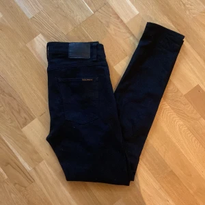 Svarta jeans från Nudie Jeans - Snygga svarta jeans från Nudie Jeans med klassisk femficksdesign. De har en diskret broderad detalj på bakfickan och en läderpatch vid midjan. Perfekta för en stilren look.