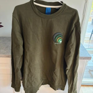 Olivgrön tröja från Scotch & Soda - Säljer en olivgrön tröja från Scotch & Soda med ett unikt broderat märke på bröstet. Tröjan har långa ärmar och en rund halsringning. Perfekt för en avslappnad stil.