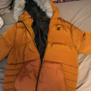 Orange SikSilk pufferjacka  - Säljer en orange pufferjacka från SikSilk med en mysig pälsdetalj på huvan. Jackan har en dragkedja framtill och är perfekt för kyligare dagar. Den har en stilren design med märkets logga på bröstet. Ny pris 1200kr, pris kan diskuteras vid snabb affär