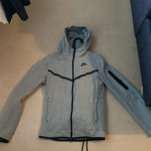 Nike tech tracksuit - Säljer en grå hoodie från Nike med dragkedja och huva. Den har en svart Nike-logga på bröstet och praktiska fickor framtill. Perfekt för en avslappnad stil. Och så ingår byxorna nike tech settet nästan aldrig använt priset kan diskuteras först till kvarn