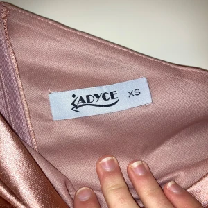 Rosa one-shoulder klänning från Adyce - Elegant rosa långklänning i satin från Adyce. Klänningen har en stilren one-shoulder design och en smickrande slits framtill. Perfekt för en student bal eller finare tillställning. Aldrig använd, endast provad 