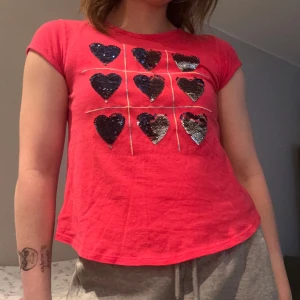 Paljett topp (GAP) - Säljer en gullig rosa t-shirt från GAP Kids med nio silver/ svarta vändbara paljetthjärtan på framsidan. T-shirten har korta ärmar och en rund halsringning. Perfekt för en glittrig touch i vardagen!