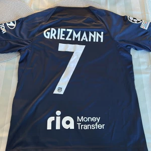Blå fotbollströja med Griezmann 7 - Snygg blå fotbollströja från Nike med Griezmanns namn och nummer 7 på ryggen. Tröjan har korta ärmar och flera sponsorloggor, inklusive Riyadh Air och UEFA Foundation. Perfekt för fotbollsfans!