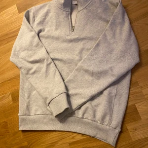 Grå tröja med dragkedja från Les Deux - Säljer en stilren grå tröja från Les Deux med halv dragkedja och broderad logga på bröstet. Perfekt för en avslappnad look. Tröjan har långa ärmar och ribbade muddar.