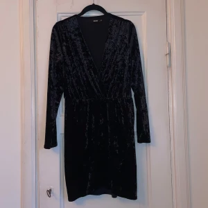 sammets klänning  - Elegant svart klänning i mjuk sammet. Den har en v-ringad design och långa ärmar, perfekt för en stilren look. Klänningen är knälång och har en omlott-effekt som ger en smickrande passform.