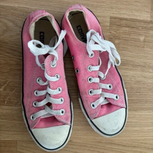 Rosa sneakers från Converse - Säljer ett par klassiska rosa Converse sneakers med vit snörning och tåhätta. Skorna har en svart rand längs sulan och är perfekta för en avslappnad stil.