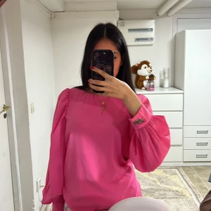 Rosa långärmad blus - Säljer en söt rosa blus med långa ärmar och rynkade detaljer vid axlarna. Blusen har en liten broderad detalj vid ärmslutet som ger en extra touch. Perfekt för en färgglad look!