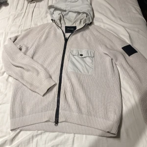 Ljusgrå stickad hoodie/cardigan från Peak Performance - Säljer en snygg ljusgrå stickad hoodie från Peak Performance. Tröjan har en dragkedja framtill och en praktisk bröstficka med knapp. Den har långa ärmar och en huva i ljusare material samt en logotyp på ärmen.Perfekt för kyliga dagar!