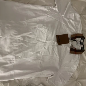 Vit t-shirt med mönstrad krage från Burberry - Säljer en stilren vit t-shirt från Burberry med en ikonisk mönstrad krage i klassiska färger. Perfekt för dig som vill ha en touch av lyx i vardagen. T-shirten har korta ärmar och en knappdetalj vid halsen. Storlek Xl men passar mer en l/m helt ny aldrig använd 