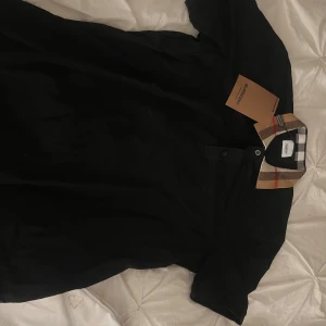 Svart t-shirt från Burberry - Snygg svart t-shirt från Burberry med klassisk rutig krage i beige, röd och vit. Perfekt för en stilren look. T-shirten har korta ärmar och knappar vid halsen. Aldrig använd ny me förpackning osv passar l/m men står större storlek på tröjan.