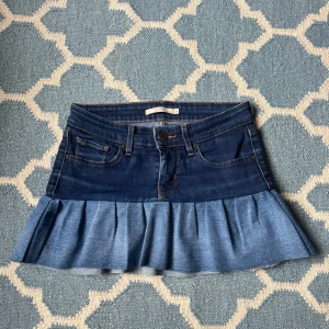 Lowwaist volangkjol, Levi's - Snygg jeanskjol från Levi's med volangdetalj i nederkant. Kjolen har en klassisk femficksdesign och är i en mörkblå denim med ljusare volang. Perfekt för en egen touch, fin under vår och sommar. Volangen går att klippa kortare om man vill. Omsydda jeans, noggrant. 