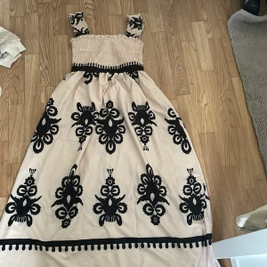Beige långklänning med svart mönster - Säljer en superfin beige långklänning med svart mönster. Klänningen har breda axelband och en smockad överdel som ger en bekväm passform. Den är perfekt för sommarens alla fester och har en unik design med stora svarta mönster som verkligen sticker ut. Klänningen är i nyskick och redo att bäras på nästa fest!