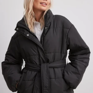 Svart pufferjacka med bälte - Säljer en svart pufferjacka som är perfekt för vintern. Jackan har en långärmad design och är utrustad med både dragkedja och knappar för extra skydd mot kylan. Den har också ett bälte i midjan för en snygg och justerbar passform. Jackan är i nyskick och redo att hålla dig varm under kalla dagar.