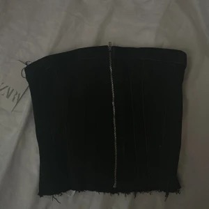 Svart tubtopp med dragkedja - Snygg svart tubtopp med dragkedja framtill. Perfekt för en utekväll eller fest. Den har en fransig kant nertill som ger en cool och avslappnad look.