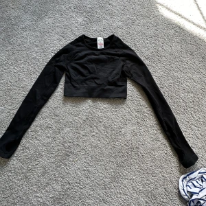 Svart croppad tröja från Gymshark - Säljer en svart croppad tröja från Gymshark. Den är perfekt för träning med sin tighta passform och långa ärmar. Materialet är en blandning av polyester och elastan, vilket gör den både bekväm och stretchig. Tröjan har en cool öppen ryggdetalj och Gymshark-loggan tryckt på baksidan. Perfekt för gymmet eller en sportig look!