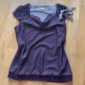 Topp - Säljer en superfin lila topp från íora. Den har en söt rosett på axeln och korta ärmar. Toppen är i ett mjukt och bekvämt material, perfekt för både vardag och fest!