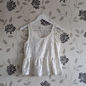 Vit spetslinne från Monki - Säljer ett spetslinne från Monki i storlek XS. Den är ärmlös och har en söt volangdetalj vid midjan som ger en feminin touch. Perfekt för sommaren eller en festlig tillställning. Linnet är i bra skick och har en tight passform. Materialet är spets vilket gör den både elegant och bekväm. Obs liten tråddragning på mitten på framsidan (se bild).
