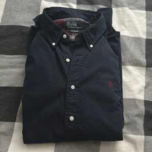 Svart skjorta från Polo Ralph Lauren - Säljer en snygg svart skjorta från Polo Ralph Lauren i storlek L. Skjortan är i nyskick och har en klassisk design med knappar och en broderad röd logga på bröstet. Perfekt för både vardag och fest! priset går o diskutera vid smidig affär